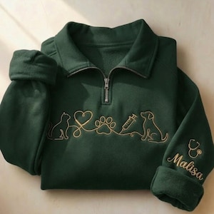 Op de afbeelding: Een donkergroene sweater met korte rits en goudkleurig borduurwerk van een kat, hart, pootafdruk, injectiespuit en hond. De rechtermouw is voorzien van een geborduurde stethoscoop en de naam Malisa in goudkleurige schrijfletters.