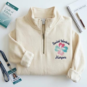 May include: A cream-colored quarter-zip sweatshirt featuring an embroidered design on the left chest. The embroidery includes the text "Social Worker" above and "Harper" below a four-leaf clover shape made of pink and teal hearts with white hand silhouettes.
