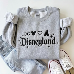 May include: A gray crewneck sweatshirt with the word "Disneyland" in black script, along with a bow, Mickey Mouse head, heart, and castle graphic. The sweatshirt is paired with light wash jeans and white sneakers.