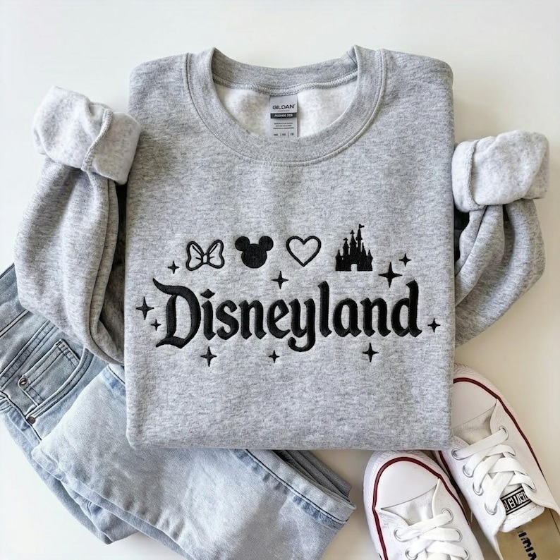 May include: A gray crewneck sweatshirt with the word "Disneyland" in black script, along with a bow, Mickey Mouse head, heart, and castle graphic. The sweatshirt is paired with light wash jeans and white sneakers.