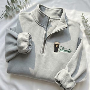 May include: A light gray sweatshirt with a quarter-zip collar and long sleeves. The sweatshirt features an embroidered design of a pint of stout and the word "Sl&aacute;inte" in green lettering.