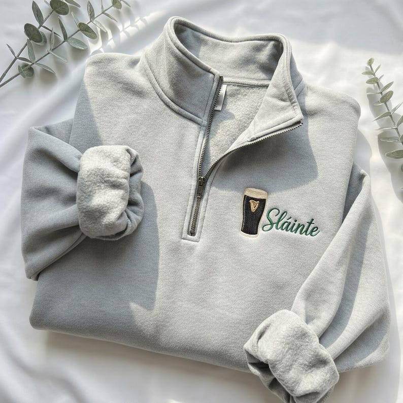 May include: A light gray sweatshirt with a quarter-zip collar and long sleeves. The sweatshirt features an embroidered design of a pint of stout and the word "Sl&aacute;inte" in green lettering.