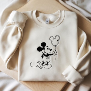 Puede incluir: Sudadera de color crema con un gráfico en blanco y negro de Mickey Mouse sosteniendo un globo con forma de Mickey Mouse. La sudadera tiene cuello redondo y mangas largas con puños acanalados.