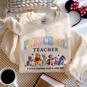 May include: A cream-colored crewneck sweatshirt featuring the word PRESCHOOL in a multi-colored gingham pattern, the word TEACHER in navy, and an illustration of Winnie the Pooh characters with the text A LITTLE KINDNESS GOES A LONG WAY.