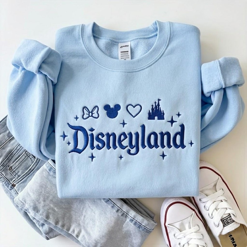 May include: Light blue sweatshirt with the word "Disneyland" embroidered in dark blue, along with Mickey Mouse ears, a bow, a heart, and a castle. The sweatshirt is paired with light wash jeans and white sneakers.