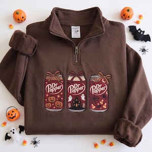 May include: A dark brown quarter-zip sweatshirt featuring an embroidered graphic of three Dr Pepper soda cans. Each can is decorated with a bow and Halloween-themed illustrations including pumpkins, a haunted house with ghosts, and spiderwebs.