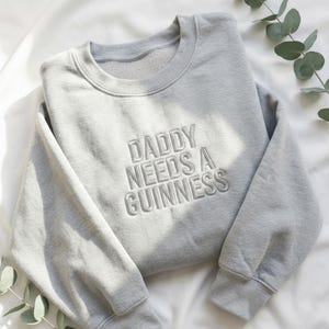 May include: A light gray sweatshirt with the embroidered text "DADDY NEEDS A GUINNESS" in a matching gray thread. The sweatshirt is folded on a white surface, with a sprig of greenery in the corner.