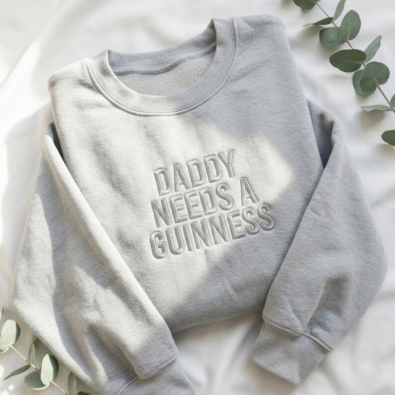 May include: A light gray sweatshirt with the embroidered text "DADDY NEEDS A GUINNESS" in a matching gray thread. The sweatshirt is folded on a white surface, with a sprig of greenery in the corner.