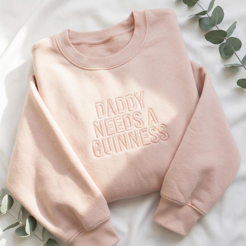 May include: A light pink sweatshirt with the embroidered text "DADDY NEEDS A GUINNESS". The crew neck sweatshirt is made of a soft material and has long sleeves. The garment is displayed on a white surface with green foliage.