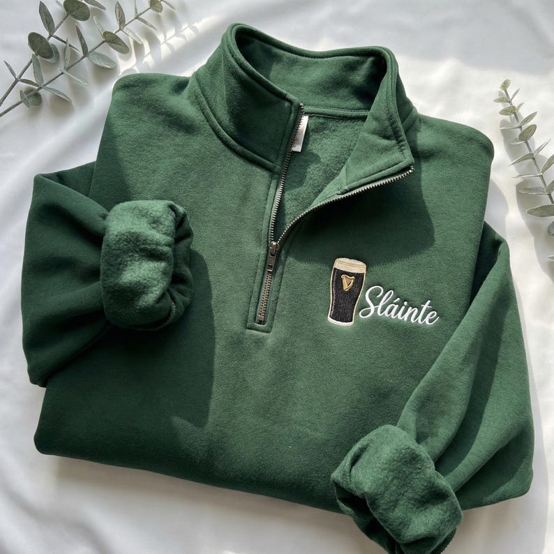 May include: A dark green quarter-zip sweatshirt with a small embroidered design of a pint of stout and the word "Sl&aacute;inte" in white script. The sweatshirt has a fleece lining and a silver zipper.