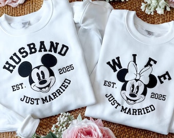 May include: Two white sweatshirts with black text and Mickey and Minnie Mouse graphics. One says "HUSBAND" with Mickey Mouse, "EST. JUST MARRIED 2025". The other says "WIFE" with Minnie Mouse, "EST. JUST MARRIED 2025".
