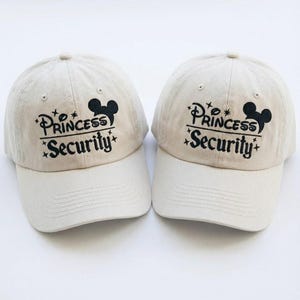 Może przedstawiać: Dwie beżowe czapki bejsbolowe z czarnym napisem i sylwetką Myszki Miki. Napis na czapkach brzmi "Princess Security" z gwiazdkami. Czapki mają zakrzywiony daszek i guzik na górze.