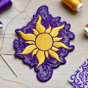 Peut inclure: Écusson brodé représentant un soleil jaune sur un fond violet en forme de dentelle. L'image comprend des bobines de fil violet et doré, une aiguille et un morceau de tissu à motifs. Idéal pour les projets de couture.