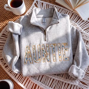 May include: A light gray quarter-zip sweatshirt featuring the word AUNTIE embroidered in a floral pattern with blue and yellow flowers on a white background, outlined in tan thread. The garment has a metal zipper and a Gildan brand label at the neck.