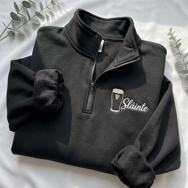 May include: Black quarter-zip sweatshirt with a white embroidered "Sl&aacute;inte" and a beer glass design. The sweatshirt has a high collar, a zipper, and cuffed sleeves. The design is on the front, near the bottom.