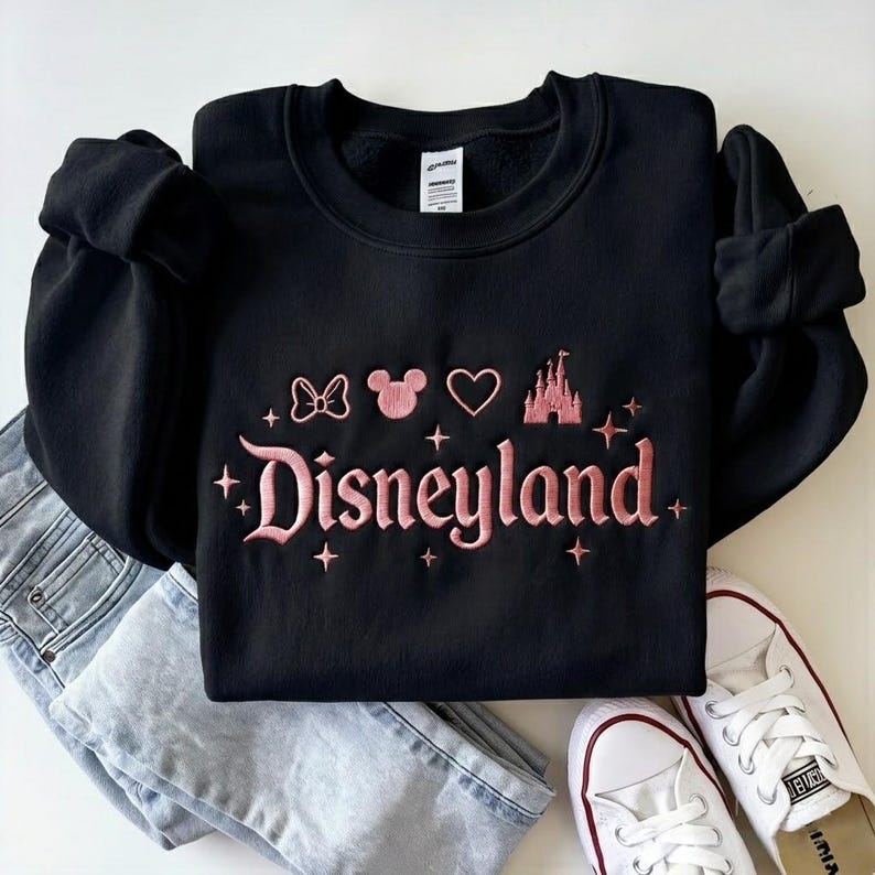May include: Black crewneck sweatshirt with the word "Disneyland" embroidered in pink script, along with a bow, Mickey Mouse head, heart, and castle. The sweatshirt is paired with light wash jeans and white sneakers.