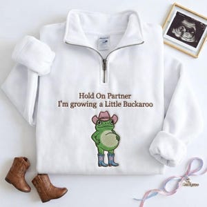 Könnte beinhalten: Ein weißes Sweatshirt mit kurzem Reißverschluss und braunem Sticktext "Hold On Partner I'm growing a Little Buckaroo" über einem gestickten grünen Frosch, der einen rosa Cowboyhut und blaue Cowboystiefel mit rosa Sternenapplikationen trägt.