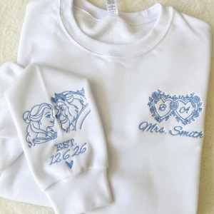 May include: A white crewneck sweatshirt featuring light blue embroidery. The chest displays two intertwined hearts with the letters B and A, and the text Mrs. Smith. The sleeve features an embroidered profile of two characters and EST. 12.6.26.