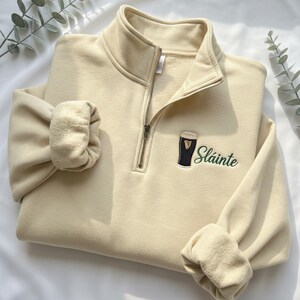 May include: Cream-colored quarter-zip sweatshirt with a black and green embroidered design of a pint of stout and the word "Sl&aacute;inte". The sweatshirt has a high collar and ribbed cuffs.