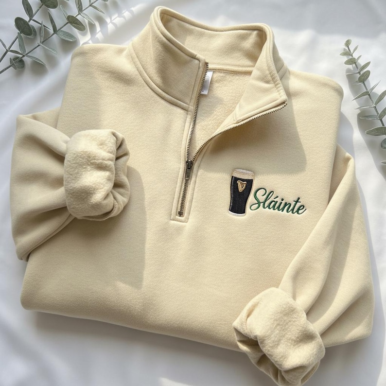 May include: Cream-colored quarter-zip sweatshirt with a black and green embroidered design of a pint of stout and the word "Sl&aacute;inte". The sweatshirt has a high collar and ribbed cuffs.