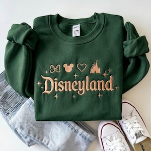 May include: A dark green sweatshirt with the word "Disneyland" embroidered in a gold color, along with Mickey Mouse icons, a castle, and stars. The sweatshirt is paired with light blue jeans and white sneakers.