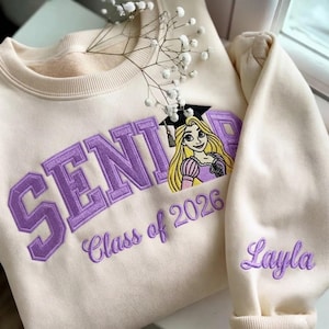 May include: A cream-coloured crewneck sweatshirt featuring purple embroidered lettering that reads SENIOR, with an embroidered illustration of a blonde character wearing a graduation cap, the text Class of 2026, and the name Layla on the sleeve.
