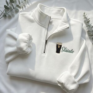 May include: A white quarter-zip sweatshirt with a black and gold embroidered beer glass and the word "Sl&aacute;inte" in green script. The sweatshirt has a high collar and a silver zipper. The sleeves are cuffed.