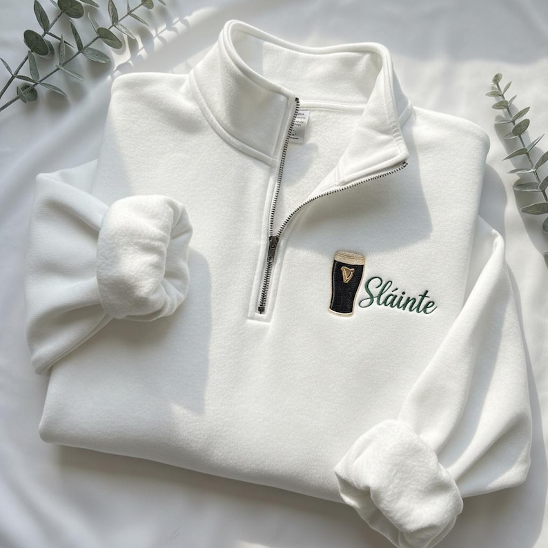 May include: A white quarter-zip sweatshirt with a black and gold embroidered beer glass and the word "Sl&aacute;inte" in green script. The sweatshirt has a high collar and a silver zipper. The sleeves are cuffed.