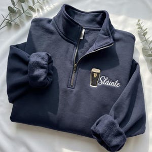 May include: Navy blue quarter-zip sweatshirt with a black and white embroidered beer glass and the word "Sl&aacute;inte" in white script. The sweatshirt has a fleece lining and ribbed cuffs.