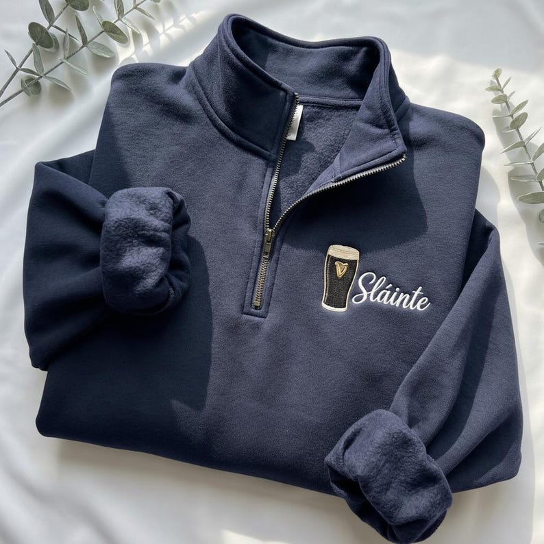 May include: Navy blue quarter-zip sweatshirt with a black and white embroidered beer glass and the word "Sl&aacute;inte" in white script. The sweatshirt has a fleece lining and ribbed cuffs.