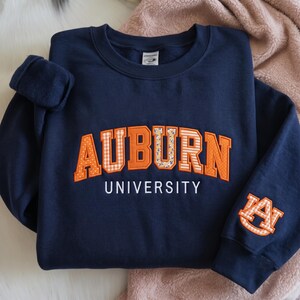 May include: A navy blue crewneck sweatshirt featuring the word AUBURN in large orange block letters with varied patterns including polka dots, gingham, and floral prints. The word UNIVERSITY is embroidered in white below, with an AU logo on the sleeve.