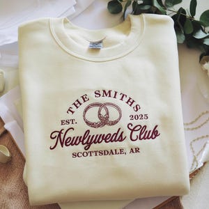 May include: Cream-coloured sweatshirt with maroon embroidered text and graphics. The design includes two wedding rings, the words "The Smiths," "Newlyweds Club," "Est. 2025," and "Scottsdale, AR."
