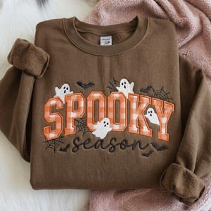 以下が含まれることがあります： オレンジと白のギンガムチェックのブロック体でSPOOKYと刺繍され、その下に黒の筆記体でseasonと刺繍された茶色のクルーネックトレーナー。白いお化け、黒いコウモリ、クモの巣の模様がデザインされています。