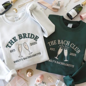 May include: White and dark green sweatshirts with embroidered text. The white sweatshirt says "THE BRIDE EST. 2025 EMILY'S BACHELORETTE" with champagne glasses. The dark green sweatshirt says "THE BACH CLUB EST. 2025 EMILY'S BACHELORETTE".