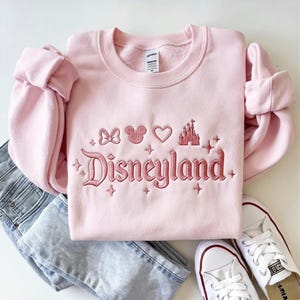 May include: A light pink sweatshirt with the word "Disneyland" embroidered in a darker pink script. Above the word are embroidered icons of a bow, Mickey Mouse ears, a heart, and a castle. The sweatshirt is paired with blue jeans and white sneakers.