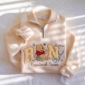 Può includere: Una felpa color crema con mezza zip, ricamata con il personaggio di Winnie the Pooh che tiene un vasetto di miele, affiancata dalle lettere R e N a quadretti gialli, uno stetoscopio, margherite e un'ape. Sotto è presente la scritta Registered Nurse.