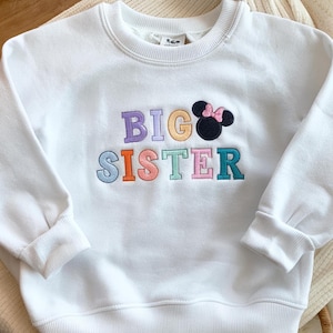 以下が含まれることがあります： カラフルな文字で「BIG SISTER」と刺繍された白いスウェットシャツ。ピンクのリボンが付いた黒いミニーマウスの頭が「BIG」の隣にあります。スウェットシャツはクルーネックで長袖です。