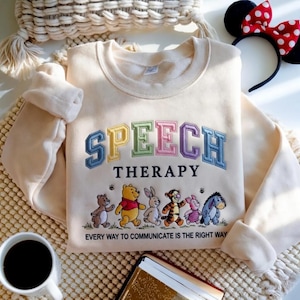 May include: A cream-coloured crewneck sweatshirt featuring the embroidered text SPEECH THERAPY in colourful block letters. Below, an embroidered illustration of Winnie the Pooh characters and the text EVERY WAY TO COMMUNICATE IS THE RIGHT WAY.