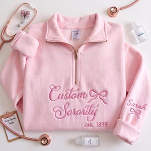 Peut inclure: Un sweat-shirt rose clair à quart de zip avec une broderie indiquant Custom Sorority est. 1895 accompagnée d'un nœud. La manche gauche porte le nom Sarah brodé avec un nœud. Le vêtement est doté d'une fermeture éclair dorée.