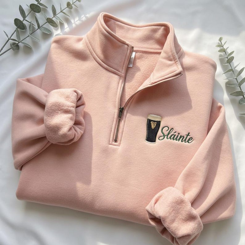 May include: A light pink sweatshirt with a quarter-zip neckline and long sleeves. The sweatshirt features an embroidered design of a pint of stout and the word "Sl&aacute;inte" in green script.