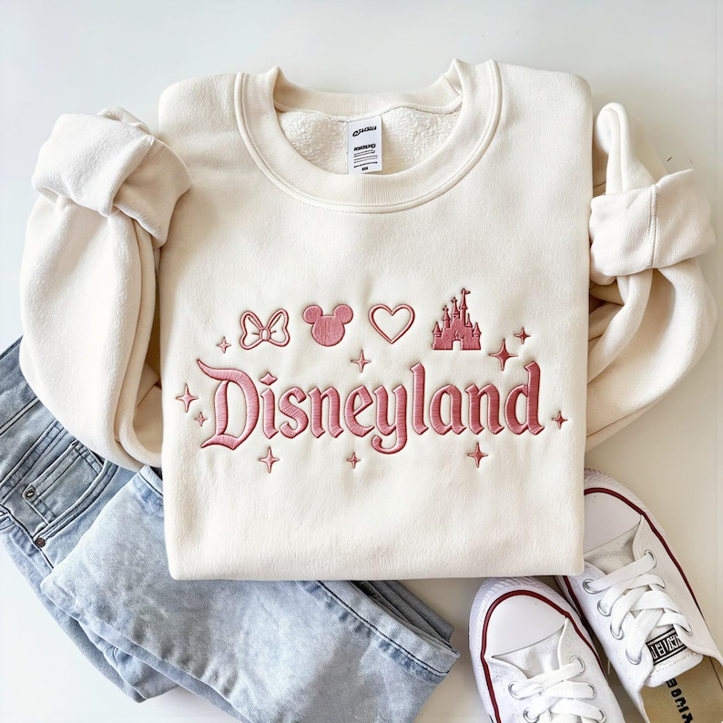 May include: Cream-colored sweatshirt with the word "Disneyland" embroidered in pink script, along with Mickey Mouse icons and a castle. The sweatshirt is paired with light-wash jeans and white sneakers.