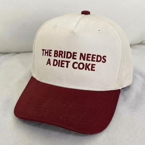 May include: A white baseball cap with a burgundy brim and top button. The text "THE BRIDE NEEDS A DIET COKE" is embroidered in burgundy on the front of the cap. This hat is a fun accessory.