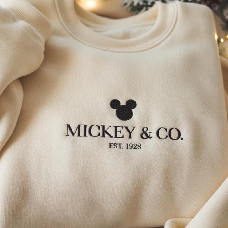 May include: Cream-colored sweatshirt with the text "MICKEY & CO. EST. 1928" in black, along with a black Mickey Mouse head graphic. The sweatshirt has a crew neck and long sleeves.