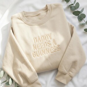 May include: A beige sweatshirt with the embroidered text "DADDY NEEDS A GUINNESS" in a matching beige thread. The sweatshirt has a crew neck and long sleeves. The fabric appears soft and comfortable.
