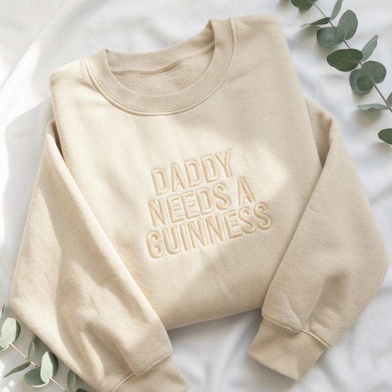May include: A beige sweatshirt with the embroidered text "DADDY NEEDS A GUINNESS" in a matching beige thread. The sweatshirt has a crew neck and long sleeves. The fabric appears soft and comfortable.