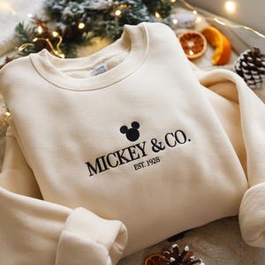 May include: Cream-colored sweatshirt with the text "MICKEY & CO. EST. 1928" in black. The design includes a Mickey Mouse silhouette above the text. The sweatshirt has a crew neck and long sleeves.