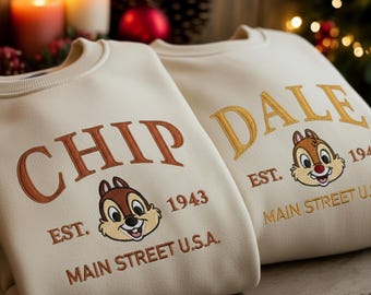 Puede incluir: Dos sudaderas de cuello redondo color crema con diseños bordados. La sudadera izquierda muestra el texto CHIP, una cara de ardilla de dibujos animados, EST. 1943 y MAIN STREET U.S.A. La derecha muestra el texto DALE, una cara de ardilla, EST. 1943 y MAIN STREET U.S.A.