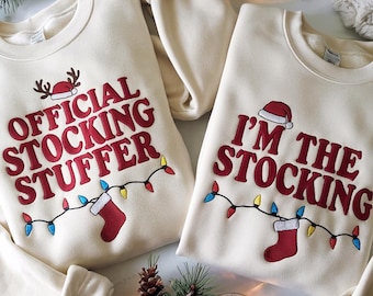 May include: Two cream-colored sweatshirts with Christmas-themed text. One says "OFFICIAL STOCKING STUFFER" and the other says "I'M THE STOCKING" in red letters. Both have a Santa hat, a red stocking, and a string of colorful lights.