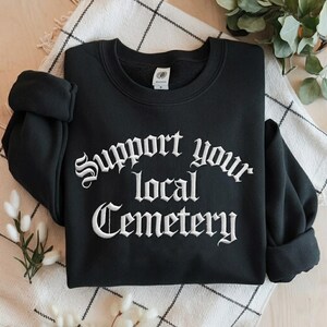 Peut inclure: Sweat-shirt noir avec l'inscription brodée en blanc "Support your local Cemetery" en lettres gothiques. Le vêtement est posé sur un tissu à carreaux noir et blanc.