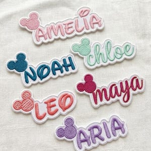 Puede incluir: Parches bordados con nombres en varios colores, incluyendo rosa, turquesa y morado. Cada parche presenta un diseño de cabeza de Mickey Mouse y un nombre: Amelia, Noah, Leo, Chloe, Maya y Aria. Los parches están sobre un fondo blanco.